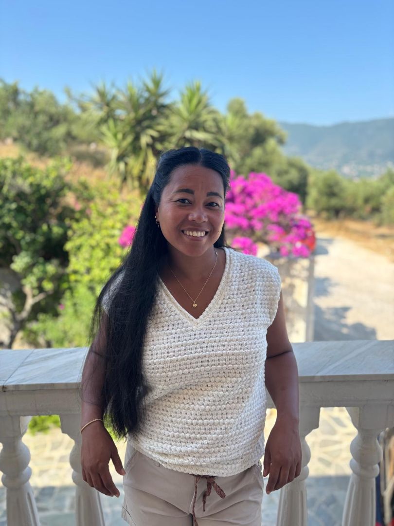 Vrouw staat buiten op een balkon met kleurrijke bloemen en groene heuvels op de achtergrond, onder een heldere blauwe lucht.