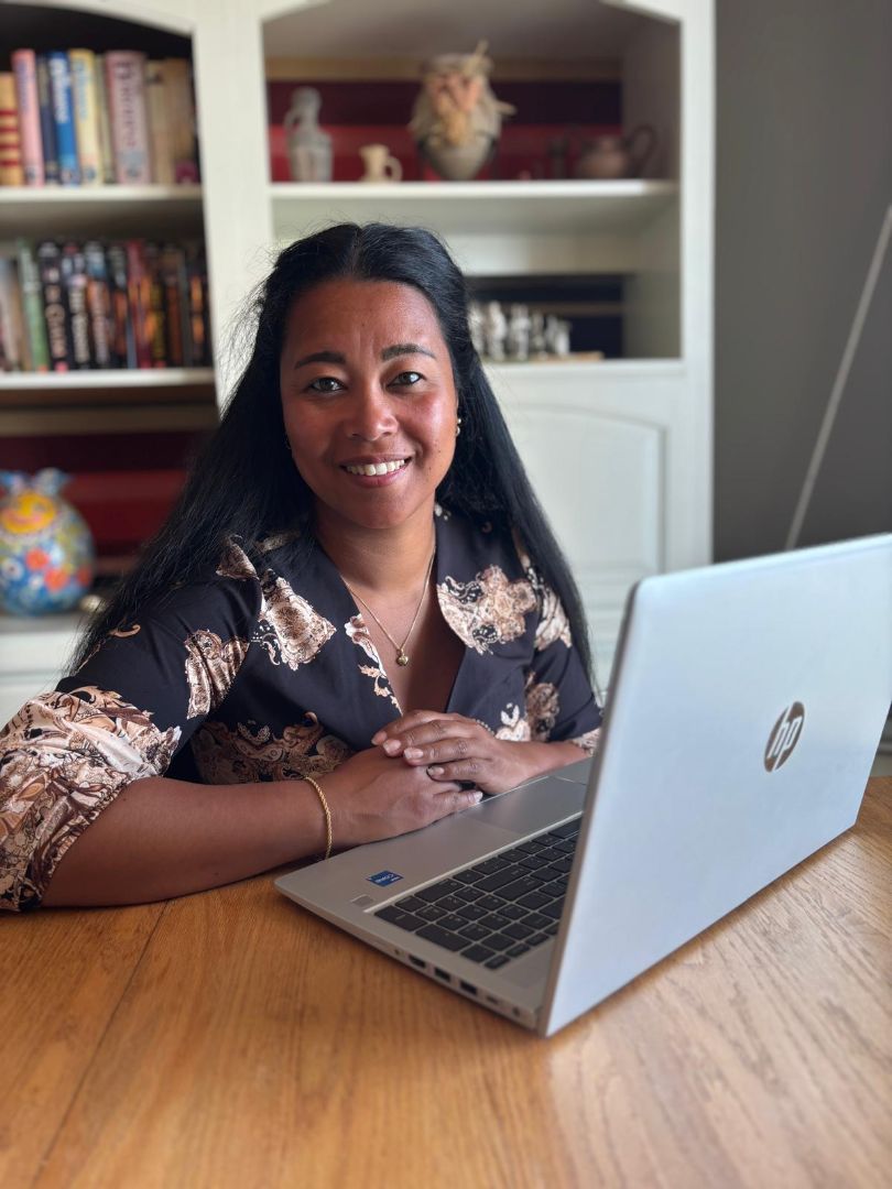 Vrouw zit aan een houten tafel met een open HP-laptop, glimlachend naar de camera. Op de achtergrond een boekenkast.