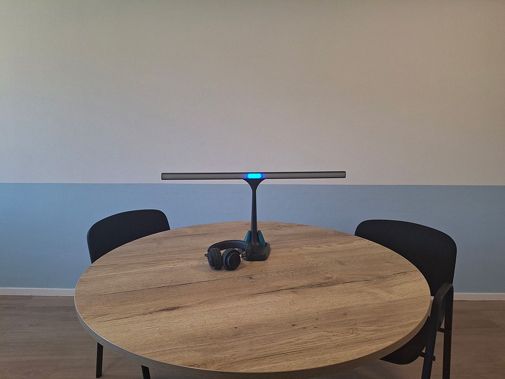 Ronde vergadertafel met twee stoelen, een modern videoconferentie-apparaat met blauwe verlichting in het midden en een koptelefoon ernaast.